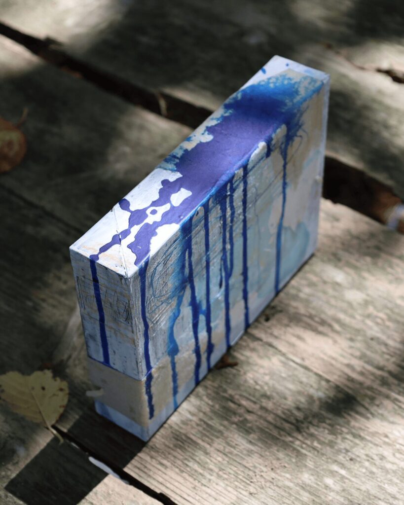"Objects, No. 1," an angled view of a sculptural mixed-media art object resting on a rustic wooden surface in dappled sunlight. This view emphasizes the object's depth, with cascades of rich blue paint flowing over the top and front faces, revealing layers of white and subtle paper collage. This piece is from the 'Objects' series by Asahi Tanaka.