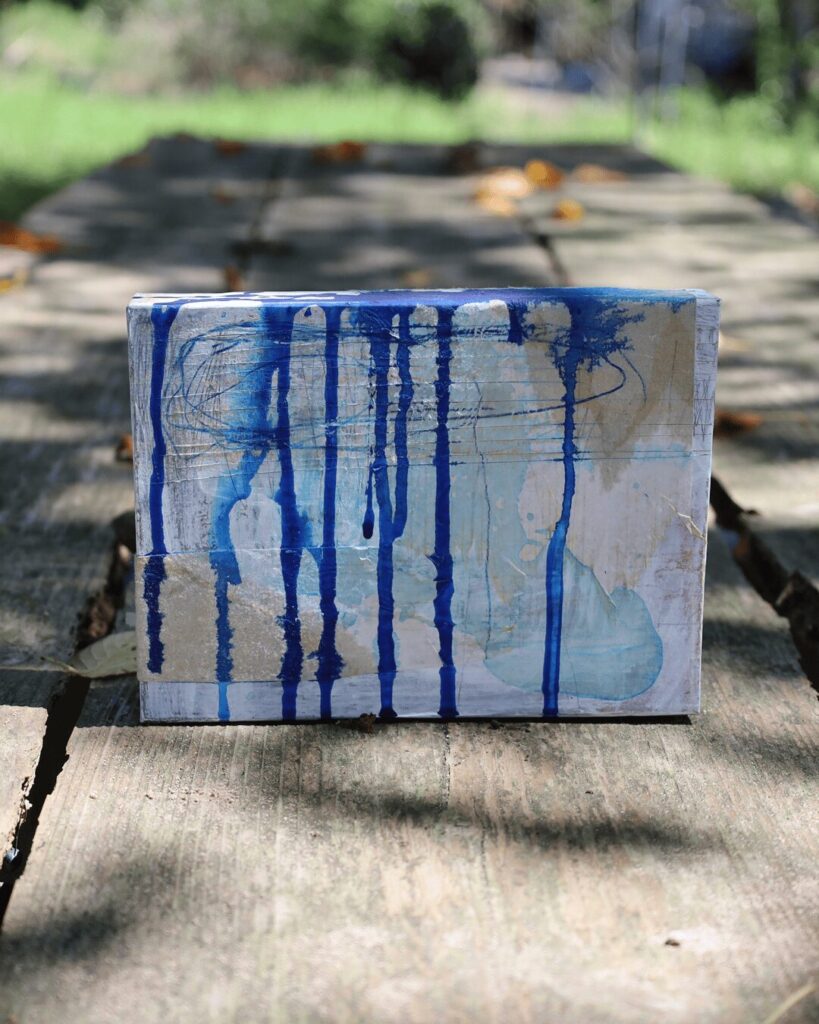 "Objects, No. 1," a frontal view of a textured abstract art object set within a natural, outdoor environment. Vertical drips of deep blue paint cascade down a weathered surface built from layers of white paint and collaged paper, showcasing the raw, honest materiality of its layered construction. This three-dimensional artwork is from the 'Objects' series by Asahi Tanaka.
