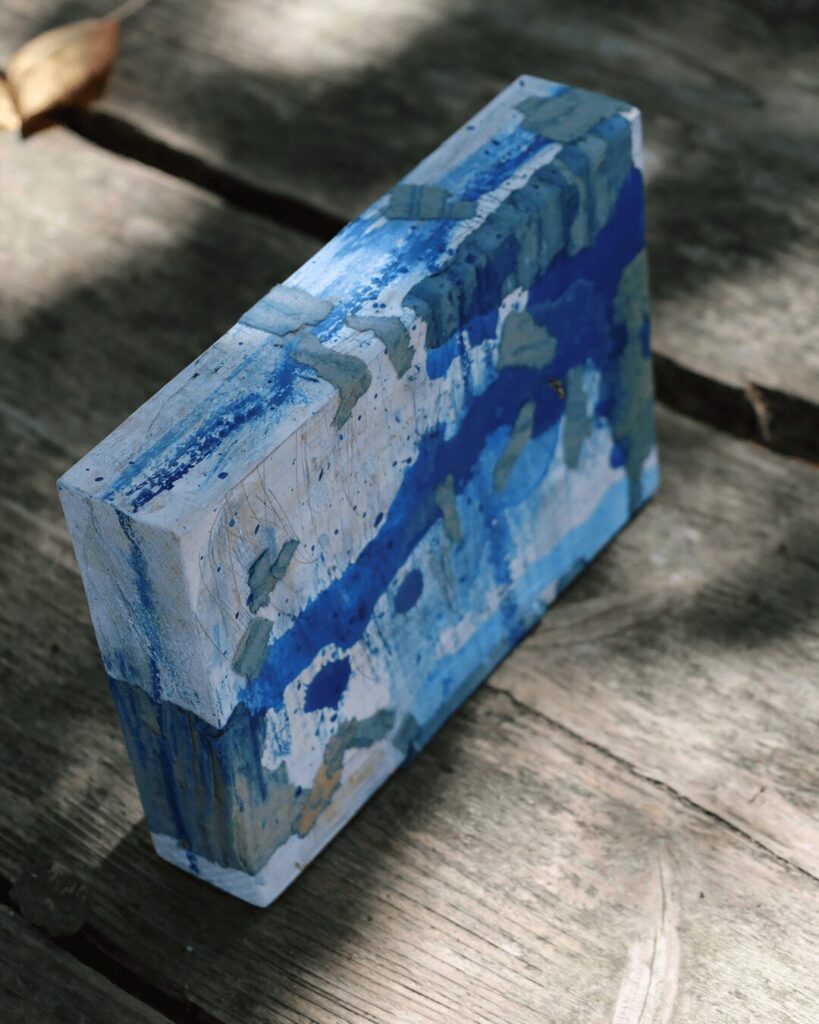 "Objects, No. 5," an angled view of a highly textured mixed-media art block, where fragments of blue-painted corrugated cardboard are collaged onto the surface. This perspective highlights the raw, dimensional quality of the collage, which overlays a more subtle background of blue drips and pencil marks. This sculptural piece is from the 'Objects' series by Asahi Tanaka.