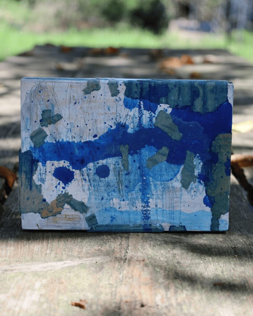 "Objects, No. 5," a frontal view of a raw, abstract collage object presented in a natural outdoor setting. The composition is dominated by collaged fragments of blue-painted cardboard, which create a rugged topography over a dynamic background of blue paint splatters and colored pencil lines. This tactile, Wabi Sabi artwork is from the 'Objects' series by Asahi Tanaka.