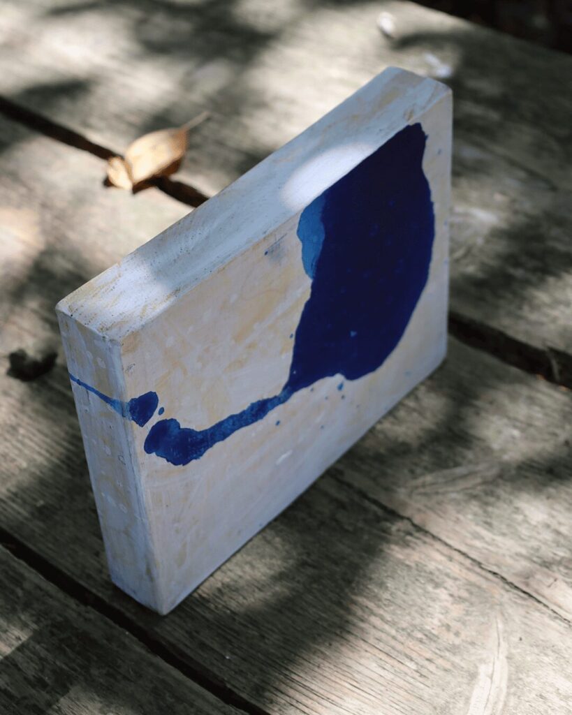 "Objects, No. 13," an angled view of a minimalist art object, showcasing the glossy, pooled texture of a single, powerful form of deep blue paint. The form's "tail" elegantly wraps around the corner of the block, emphasizing the artwork's sculptural quality against its weathered, off-white surface. This Japandi style piece is from the 'Objects' series by Asahi Tanaka.
