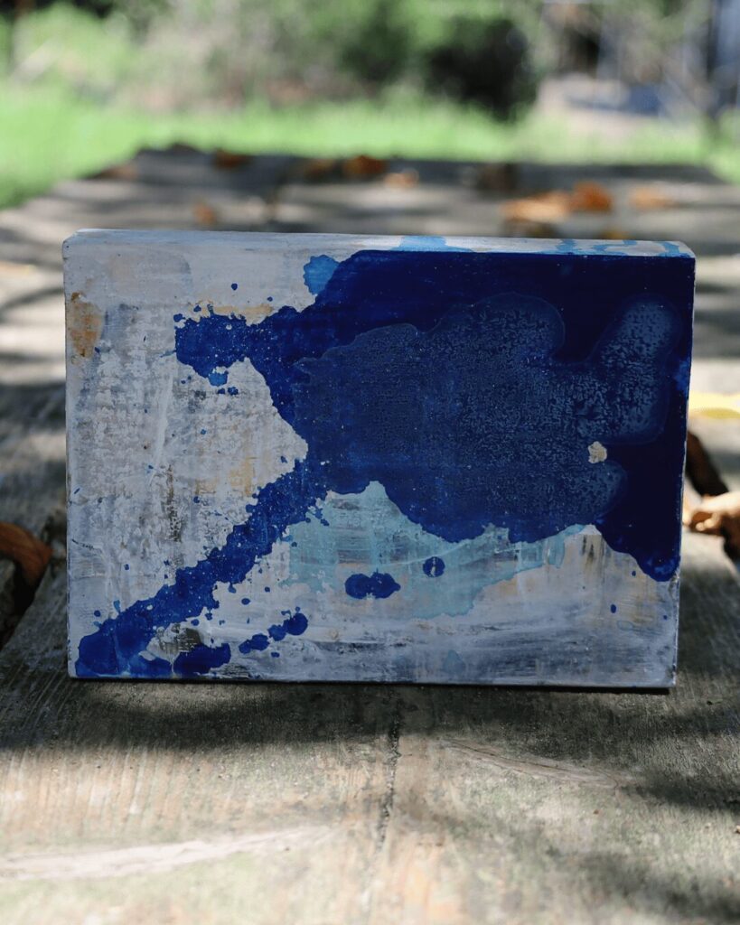 "Objects, No. 12," a serene frontal view of a Japandi style abstract art block presented in a natural outdoor setting. The composition features several large, organic forms of deep and light blue paint that have pooled and spread across a subtly textured, off-white background, creating a quiet and contemplative visual field. This piece of contemporary art is from the 'Objects' series by Asahi Tanaka.