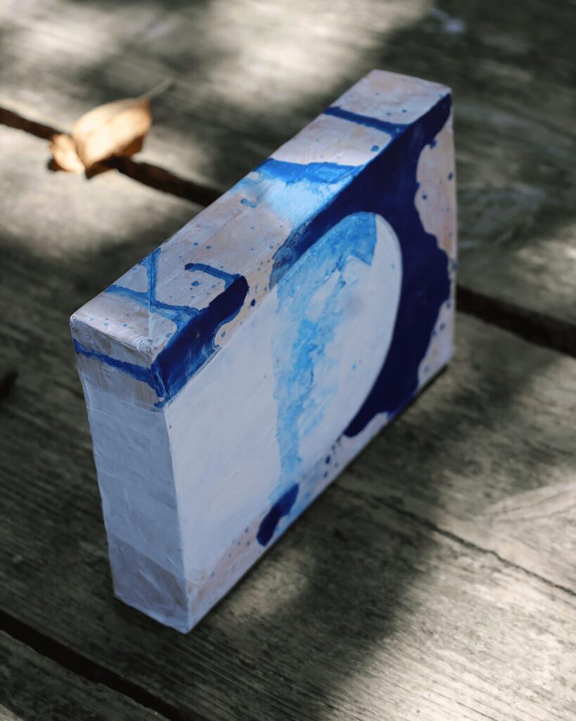 "Objects, No. 11," an angled view of a sculptural art block resting in dappled sunlight. This perspective shows how a bold, clean white form is framed by a deep navy blue layer, with both shapes wrapping around the object's corner, emphasizing its solid, three-dimensional presence. This piece is from the 'Objects' series by Asahi Tanaka.