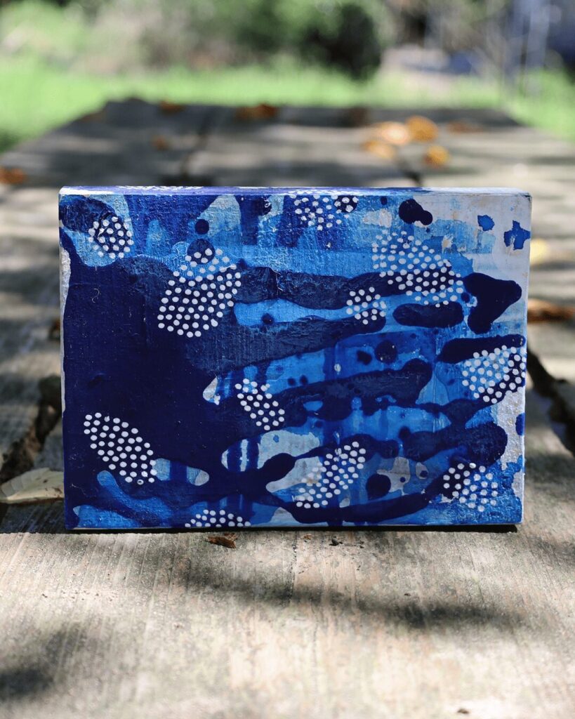 "Objects, No. 2," a captivating frontal view of a contemporary art object placed on a weathered wooden plank. Mesmerizing clusters of white dots float and gather across a deep, fluid background of layered blue paint, creating a beautiful interplay between precise order and organic chaos. This piece is from the 'Objects' series by Asahi Tanaka.