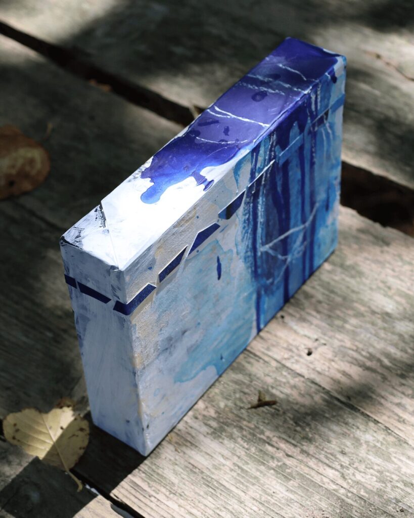 "Objects, No. 6," an angled, three-quarter view of a complex mixed-media art block, showing how collaged chips of blue paper and delicate white crayon marks create a structured layer over a fluid background of blue drips. This perspective reveals the object's rich, multi-layered history. This sculptural piece is from the 'Objects' series by Asahi Tanaka.
