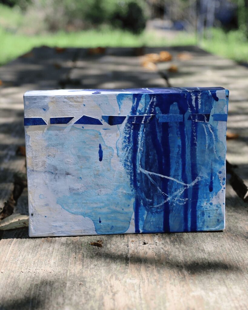 "Objects, No. 6," a frontal view of a multi-layered contemporary art object resting on a weathered wooden plank. The composition features delicate white crayon lines drawn over a dynamic collage of blue paper chips and cascading blue paint, creating a dialogue between intentional marks and accidental forms. This minimalist, textured artwork is from the 'Objects' series by Asahi Tanaka.
