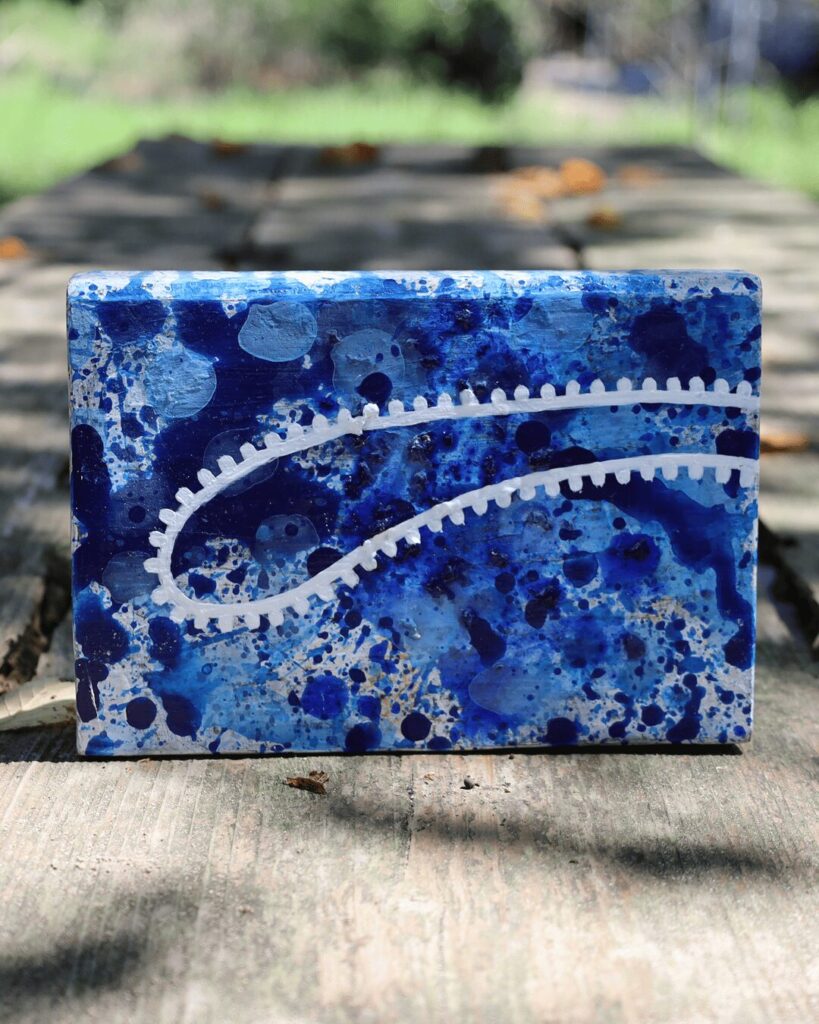 "Objects, No. 7," a powerful frontal view of a contemporary art block presented in natural sunlight. The composition is defined by a bold, zipper-like white line that carves a deliberate path through a dynamic, multi-layered blue background, creating a dialogue between precise control and organic energy. This textured artwork is from the 'Objects' series by Asahi Tanaka.