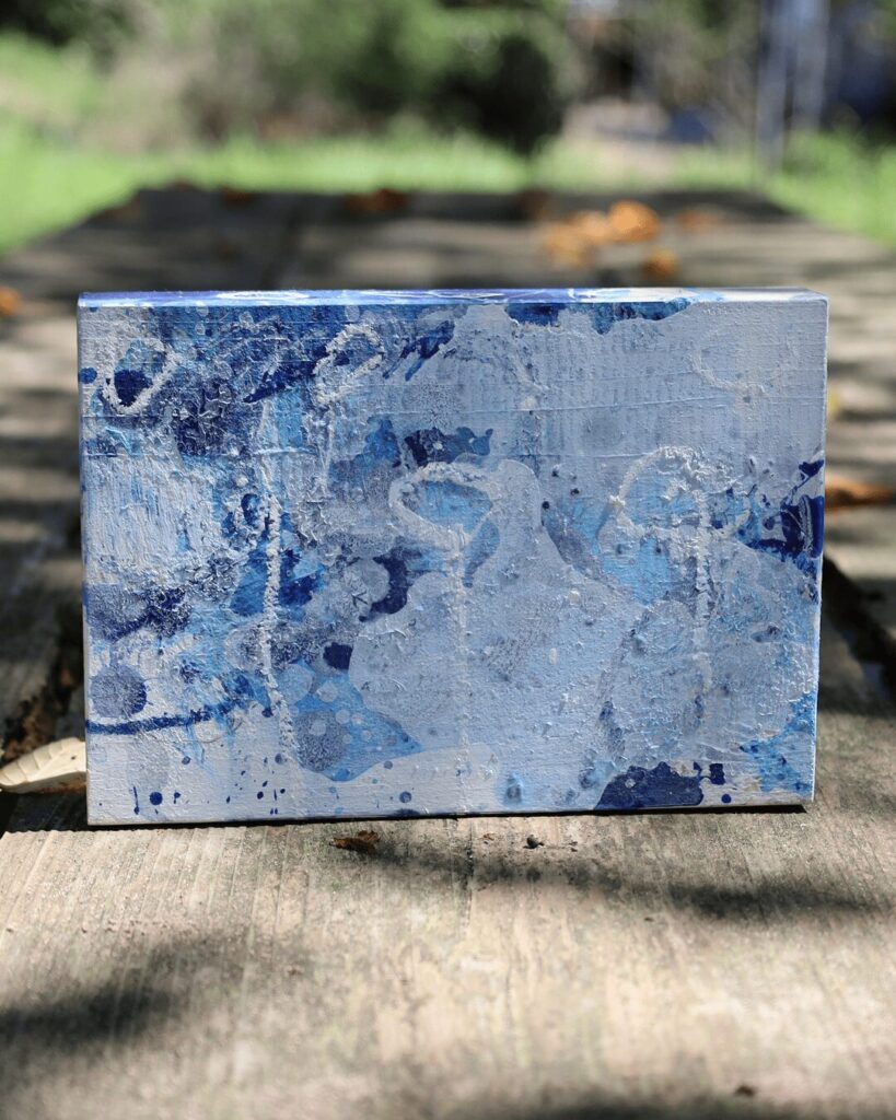 "Objects, No. 4," a frontal view of a layered abstract art block set against a rustic, outdoor backdrop. The composition features a distinct layer of hand-drawn white crayon marks, including circles and lines, superimposed over a fluid, multi-layered background of blue and white paint. This creates a direct dialogue between controlled, graphic mark-making and the organic, fluid forms of the paint beneath. This minimalist, textured artwork is from the 'Objects' series by Asahi Tanaka.
