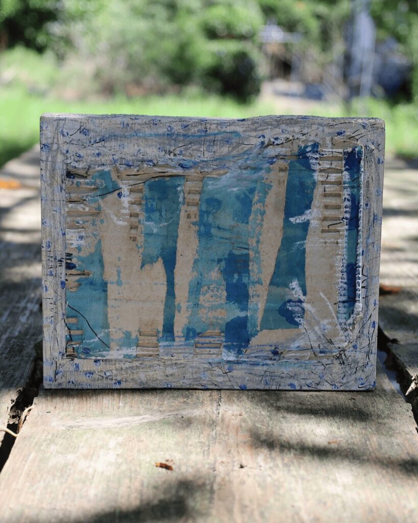 "Objects, No. 10," a powerful frontal view of a deconstructed mixed-media collage resting in dappled sunlight. The artwork features a heavily distressed surface where layers of blue paint have been peeled back to reveal the raw, corrugated texture of the underlying cardboard, creating a powerful dialogue between the finished, painted surface and the raw materials beneath. This sculptural artwork is from the 'Objects' series by Asahi Tanaka.