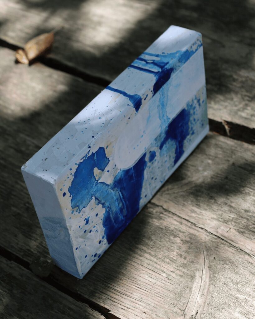 "Objects, No. 9," an angled view of a layered abstract art block resting in natural sunlight. This perspective highlights how a smooth, solid white shape has been superimposed over a dynamic background of blue splatters, emphasizing the object's three-dimensional form and the interplay between chaos and control. This piece is from the 'Objects' series by Asahi Tanaka.
