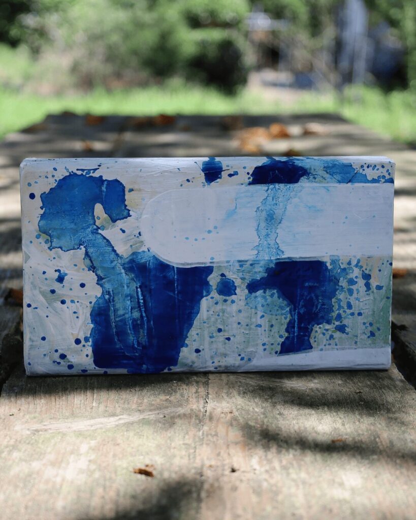 "Objects, No. 9," a minimalist frontal view of a contemporary art object set on a rustic wooden surface. The composition features a bold, clean white form that creates a window of calm, partially concealing and editing an energetic underlayer of deep blue paint splatters. This textured artwork is from the 'Objects' series by Asahi Tanaka.