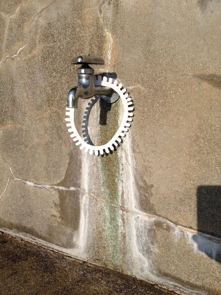A photographic artwork from the 'Encounters' series by Asahi Tanaka. This photograph documents a site-specific art intervention where a single white, gear-like ring is hung around the base of a metal water faucet. The faucet is fixed to a weathered concrete wall that shows cracks and mineral stains, and the scene is lit by direct sunlight, casting a sharp shadow of the ring and faucet.