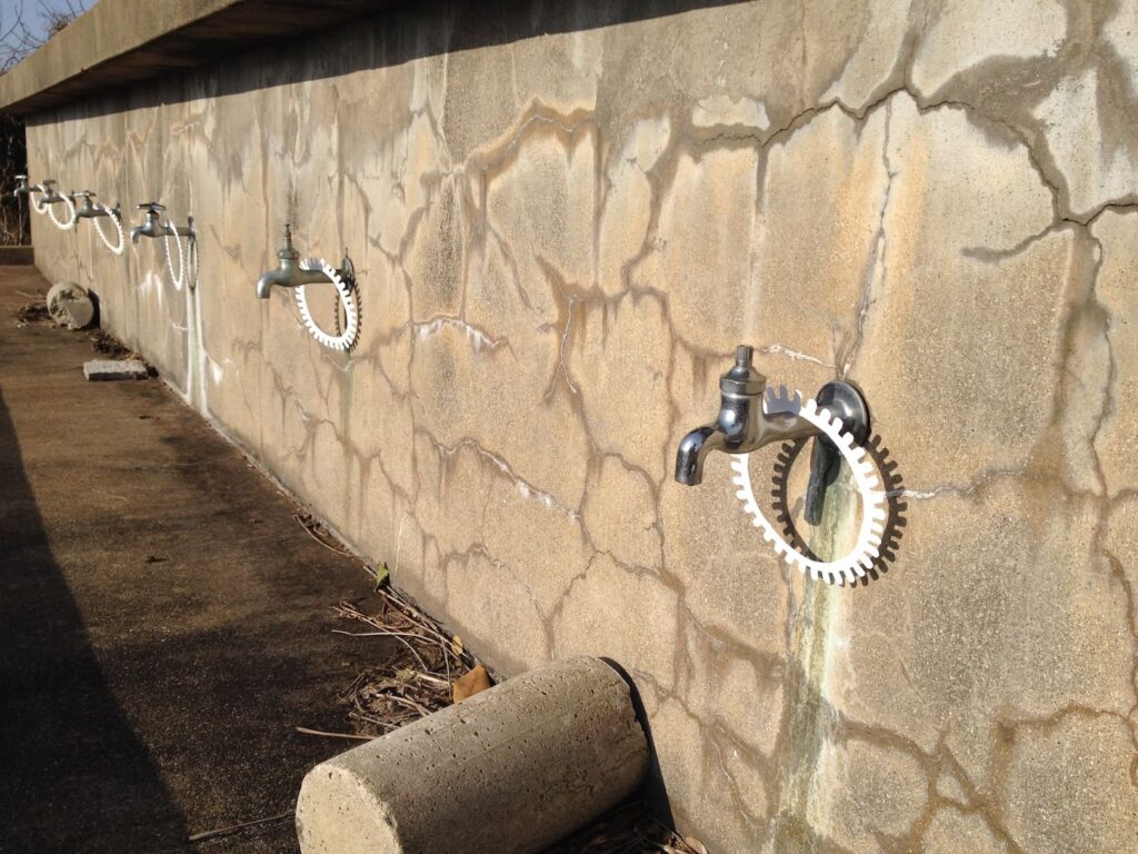 A photographic artwork from the 'Encounters' series by Asahi Tanaka. This is an angled perspective photograph of a site-specific art intervention at an outdoor water facility. Several white, gear-like rings are hung around a row of metal faucets that are mounted on a long, cracked concrete wall. The repeating placement of the rings on the functional objects creates a rhythmic, graphic pattern along the wall.