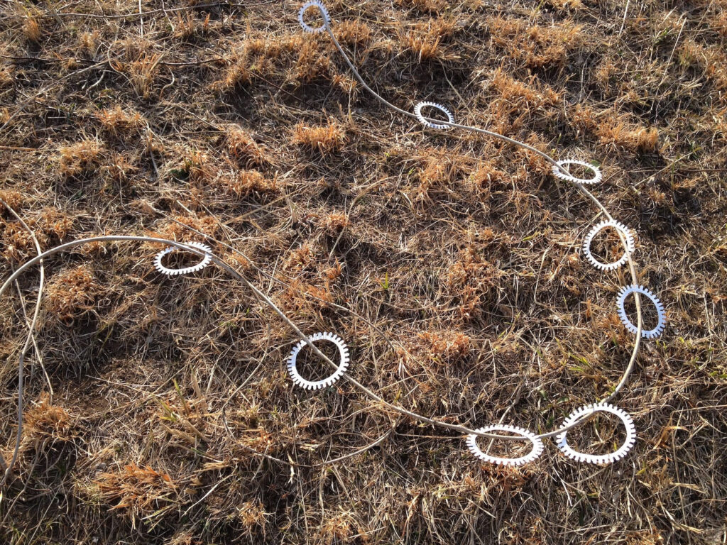 A photographic artwork from the 'Encounters' series by Asahi Tanaka. This photograph documents an art intervention in a field of dry, brown grass. Multiple white, gear-like rings have been threaded onto a long, thin vine, which is then laid out on the ground in a meandering, circular path. This creates a deliberate arrangement of the repeating geometric forms within the natural landscape.