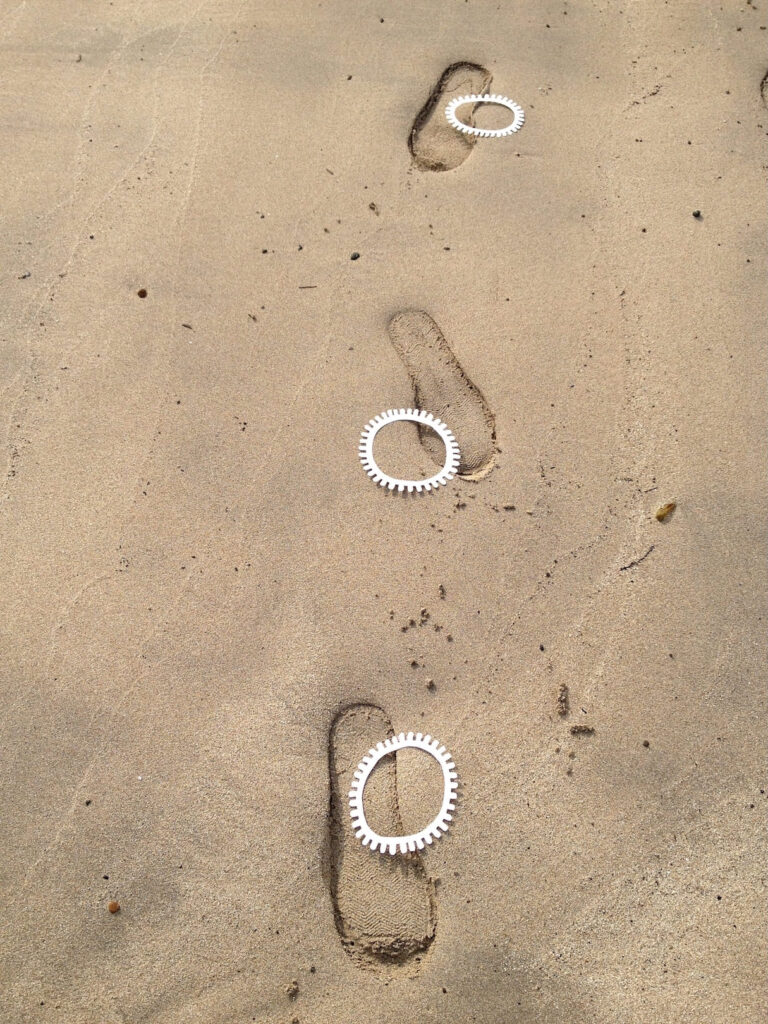 A photographic artwork from the 'Encounters' series by Asahi Tanaka. This photograph captures a site-specific art intervention on a sandy beach. Three white, gear-like rings are individually placed inside three human footprints that form a single line pressed into the damp sand. The composition documents the direct interaction between the repeating manufactured objects and the temporary imprints of a human presence.