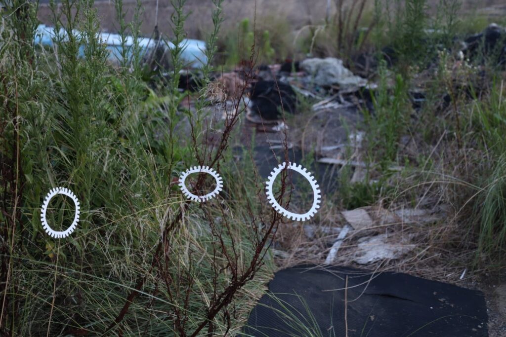 A photographic artwork from the 'Encounters' series by Asahi Tanaka. This photograph documents an art intervention in an overgrown, untended area. Three white, gear-like rings are suspended from the thin, bare stalks of a plant, positioned against a background of dense green and brown weeds and discarded debris. The composition places the clean, geometric forms of the rings within a chaotic, wild environment.