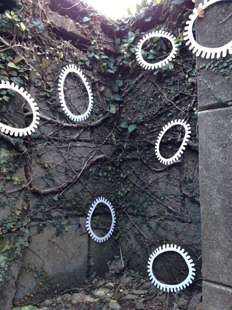 A photographic artwork from the 'Encounters' series by Asahi Tanaka. This is an angled photograph of an art intervention on a concrete block wall partially covered in a network of green, creeping vines. Seven white, gear-like rings are arranged at various angles on the textured surface, creating a direct juxtaposition between the repeating man-made objects and the organic growth.