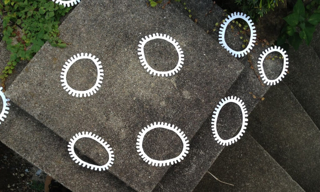 A photographic artwork from the 'Encounters' series by Asahi Tanaka. This is a high-angle photograph of a site-specific art intervention on a set of outdoor concrete stairs and a landing. Multiple white, gear-like rings are scattered across the different levels of the weathered, grey concrete, with some green foliage visible at the edge of the frame. The arrangement creates a path of repeating geometric forms that follow the angular structure of the steps.