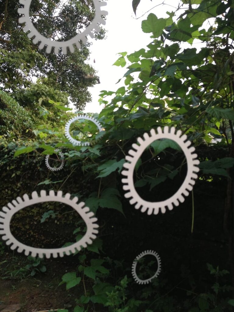 A photographic artwork from the 'Encounters' series by Asahi Tanaka. This photograph documents an art intervention where multiple white, gear-like rings are suspended from spider webs amidst the green foliage of trees and bushes. The rings seem to float at various depths and angles in the air, creating a composition that places the geometric objects within the complex, organic structure of the plant life.