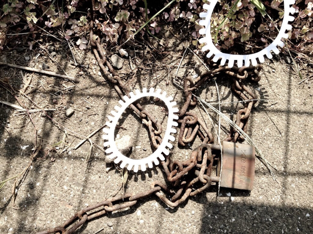 A photographic artwork from the 'Encounters' series by Asahi Tanaka. This photograph documents a site-specific art intervention where two white, gear-like rings are placed on and amongst a length of heavy, rusted chain and a padlock lying on the ground. The entire scene is overlaid with the sharp, diagonal shadow cast by a fence, adding a layer of geometric pattern to the composition of industrial objects and manufactured forms.