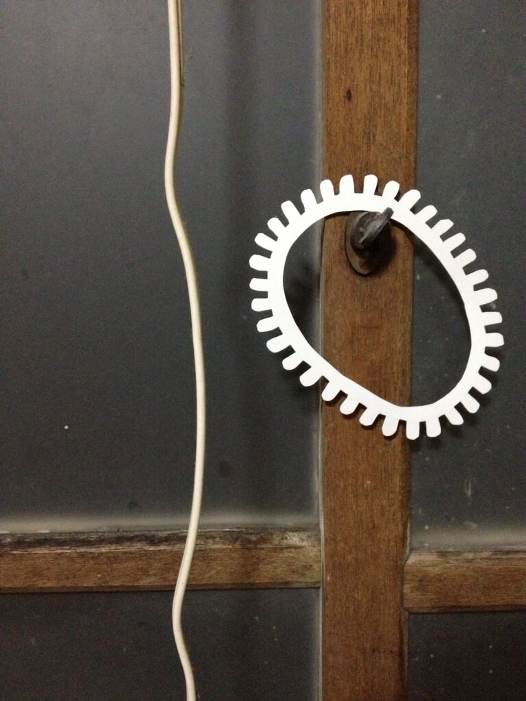 A photographic artwork from the 'Encounters' series by Asahi Tanaka. This close-up photograph captures a single white, gear-like ring hung on a small metal latch on the vertical wooden frame of an old window. The composition juxtaposes the clean, geometric form of the ring with the aged textures of the wood and frosted glass, and the soft curve of a white electrical cord to the left.