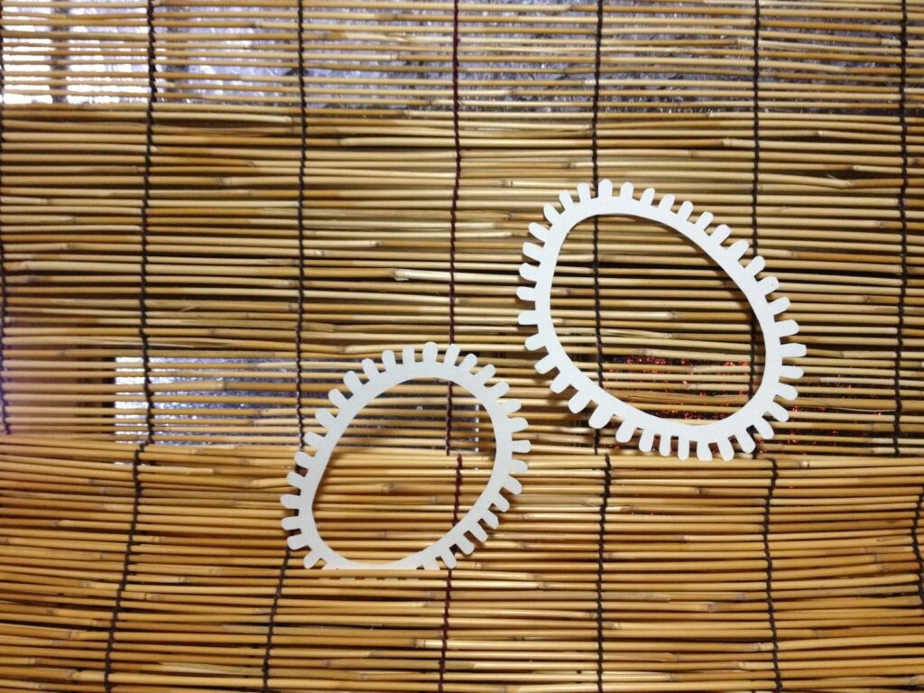 A photographic artwork from the 'Encounters' series by Asahi Tanaka. This photograph captures two white, gear-like rings of different data-sizes hung on the surface of a traditional Japanese sudare (bamboo blind). The composition juxtaposes the clean, manufactured forms of the rings with the natural texture and rhythmic, grid-like pattern of the woven bamboo.