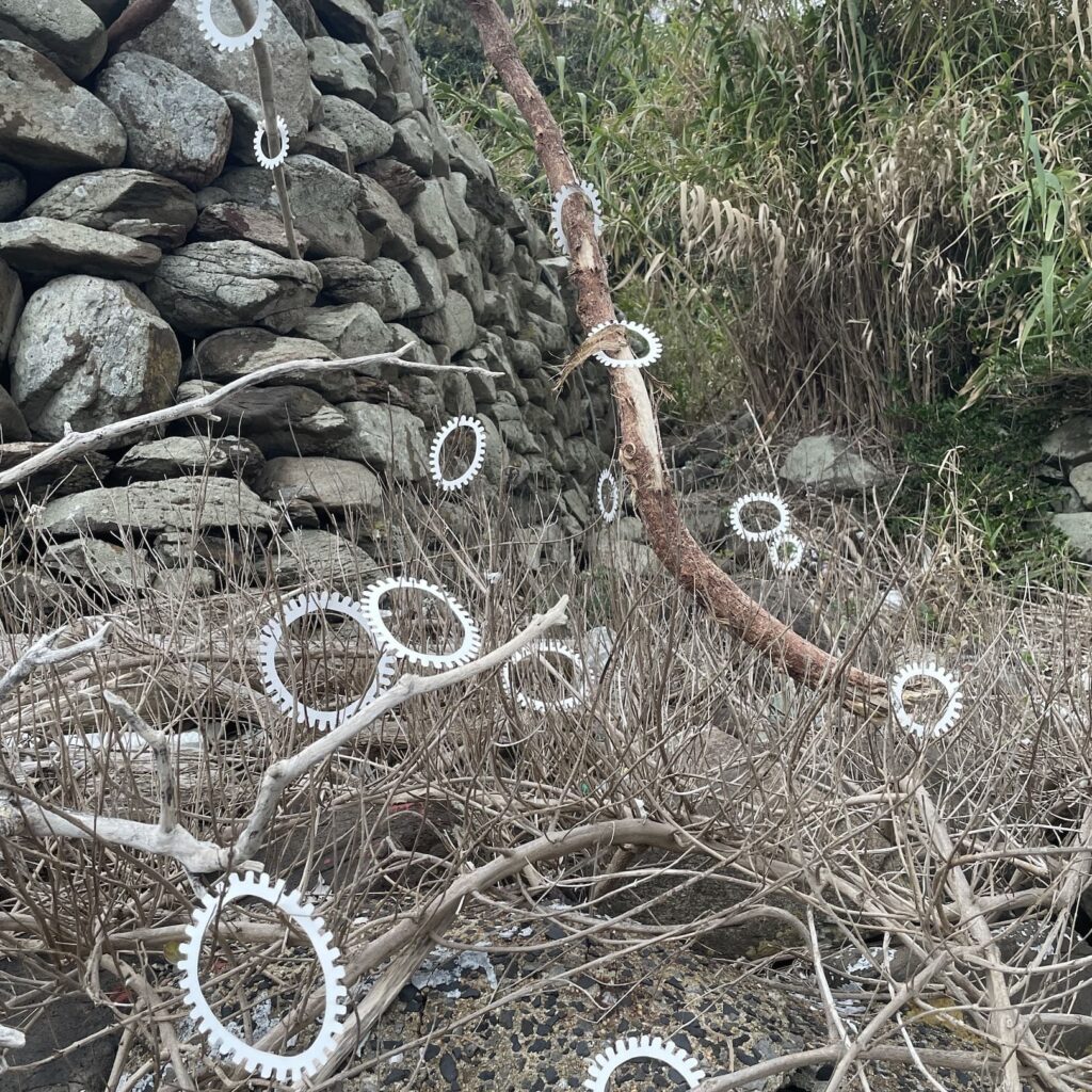 A photographic artwork from the 'Encounters' series by Asahi Tanaka. This photograph documents a site-specific art intervention in front of a stacked stone wall at the seaside. A large number of white, gear-like rings are scattered on and within a chaotic mass of dry, tangled driftwood and branches, integrating the repeating geometric forms into the complex texture of the natural debris.