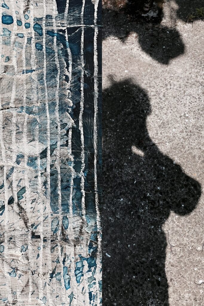 "A digital collage from the 'Correspondences' series by Asahi Tanaka. This artwork is a diptych, split vertically. The left section is a close-up detail of a heavily textured abstract painting, with a pattern of scraped, vertical white and blue crayon lines on a dark, multi-layered base. The right section is a high-contrast photograph of the artist's own shadow, along with the shadow of a plant, cast onto a rough concrete surface in direct sunlight. The composition juxtaposes the abstract, man-made texture of the painting with the ephemeral, figurative form of the shadow, finding a visual correspondence in the shared verticality and the high-contrast textures of both images. This experimental digital artwork is from the 'Correspondences' collection by Asahi Tanaka."